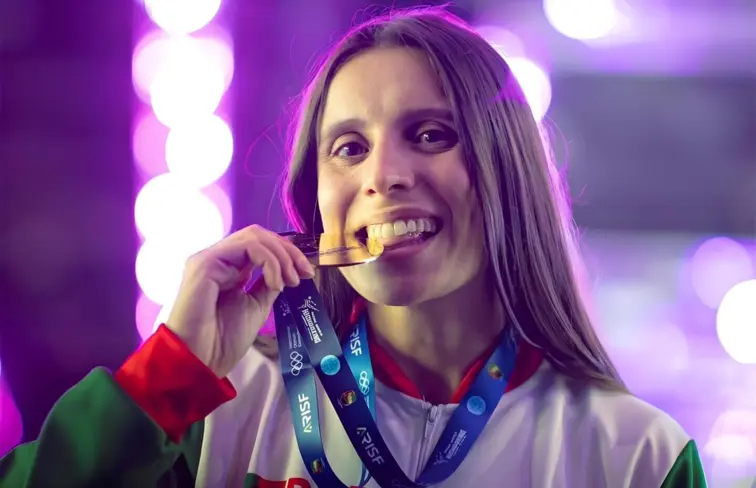 Fotografia de Sofia Oliveira, jovem de cabelo castanho esticado, a trincar a medalha de ouro, com a mão direita, enquanto sorri.
