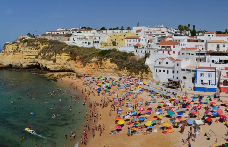 Praia de Carvoeiro, no Algarve cheia de pessoas, onde se vê a costa, com rochedos e casas num dia solarengo.