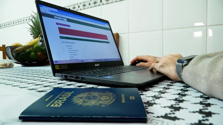 Um computador aberto no portal de renovações de autorizações de residência da AIMA, com uma mão no rato e outra em cima do portátil, ao lado está um passaporte do Brasil.